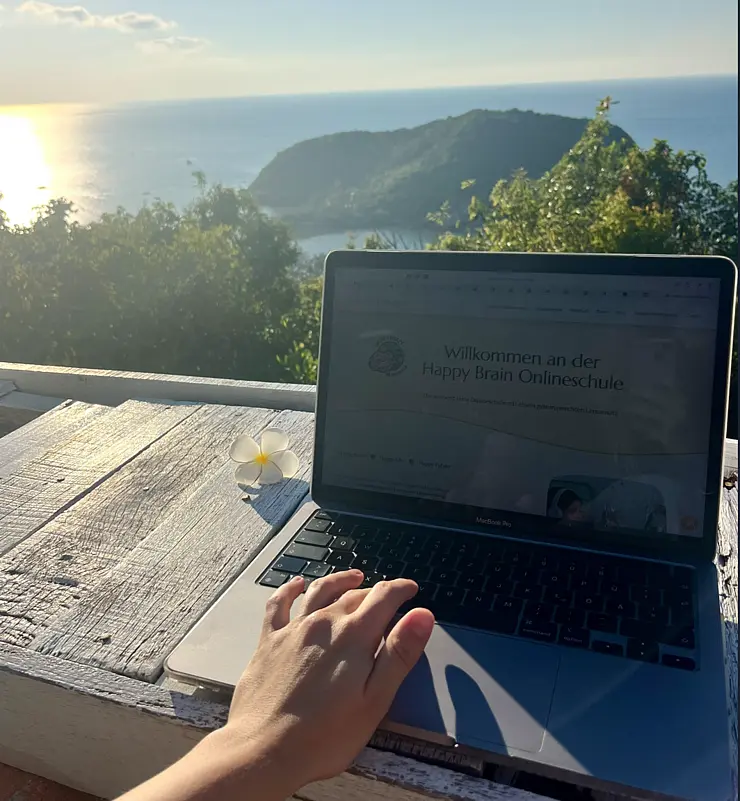 Laptop auf einem Tisch, dahinter ein Sonnenuntergang über dem Meer