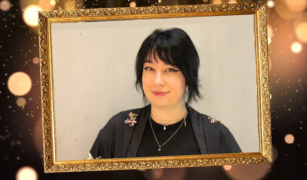 Portätbild von Lilu Kruspe - weiße junge, geschminkte Frau mit dunklen langen Haaren, in Kamera lächelnd, sie trägt ein schwarzes japanisches Gewand, um sie ein Goldrahmen und Galaglitzer