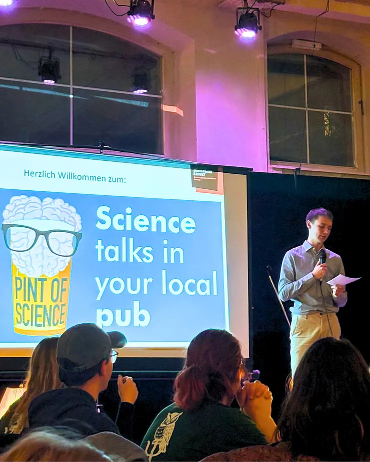 Foto einer Veranstaltung: verschiedene Personen von hinten zu sehen, im Dunkeln, auf einer Bühne Leinwand mit Gehirn im Bierglas "Science tals in your local pub" und rechts steht Jesse mit Zetteln und Mikrofon in der Hand