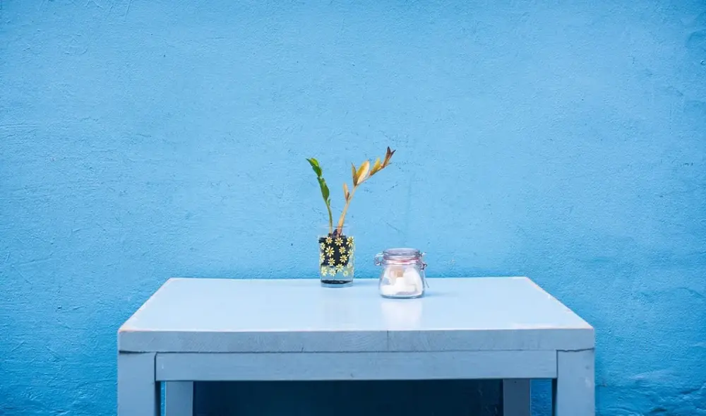 Symbolbild Raumgestaltung (ein blauer Tisch steht vor einer blauen Wand, auf dem Tisch steht eine Vase mit Pflanzenzweigen und ein Vorratsglas)