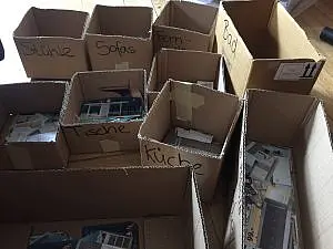Für den Workshop "Wohnen" für die Fachtagung "Leben. So, wie ich will!" sind Kartons mit Räumen und Möbeln wie Küche, Bad, Tische, Stühle, Sofas beschriftet. Darin befinden sich verschiedene Materialien für diese Räume/Möbel.
