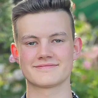 Porträtfoto von Jona, kurzes dunkelblondes Haar, leichtes Lächeln, im Hintergrund blühende Pflanzen unscharf