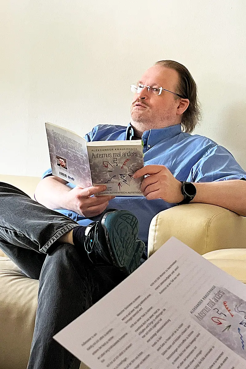Ein Mann mit mittellangen braunen haaren und Brille im blauen Hemd spricht und hält ein Buch in seinen Händen