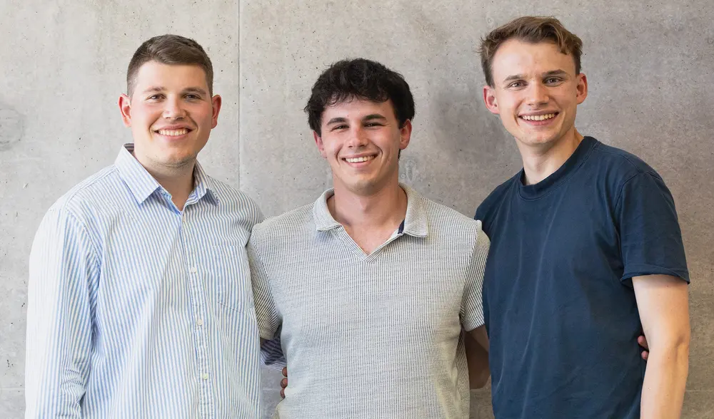 Gruppenbild mit drei jungen Männern - Elias, Kamil und Max, sich leicht umarmend, lächelnd in Kamera, vor grauem Hintergrund