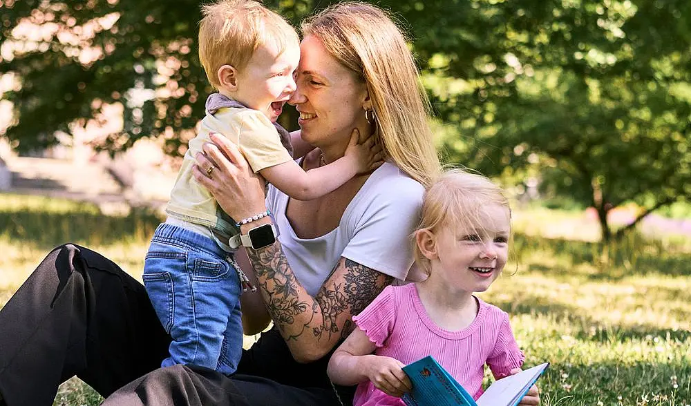 Charly, weiße Frau mit blonden langen haaren, hält ihren Sohn im Arm, ihre Tochter sitzt davor und freut sich, ein Buch haltend, alle sitzen auf einer Wiese