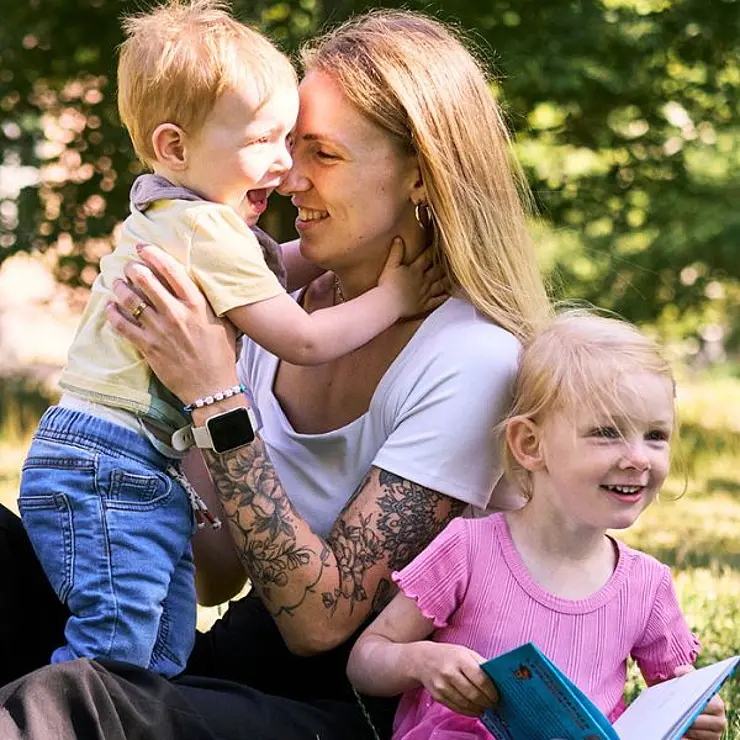 Charly, weiße Frau mit blonden langen haaren, hält ihren Sohn im Arm, ihre Tochter sitzt davor und freut sich, ein Buch haltend, alle sitzen auf einer Wiese