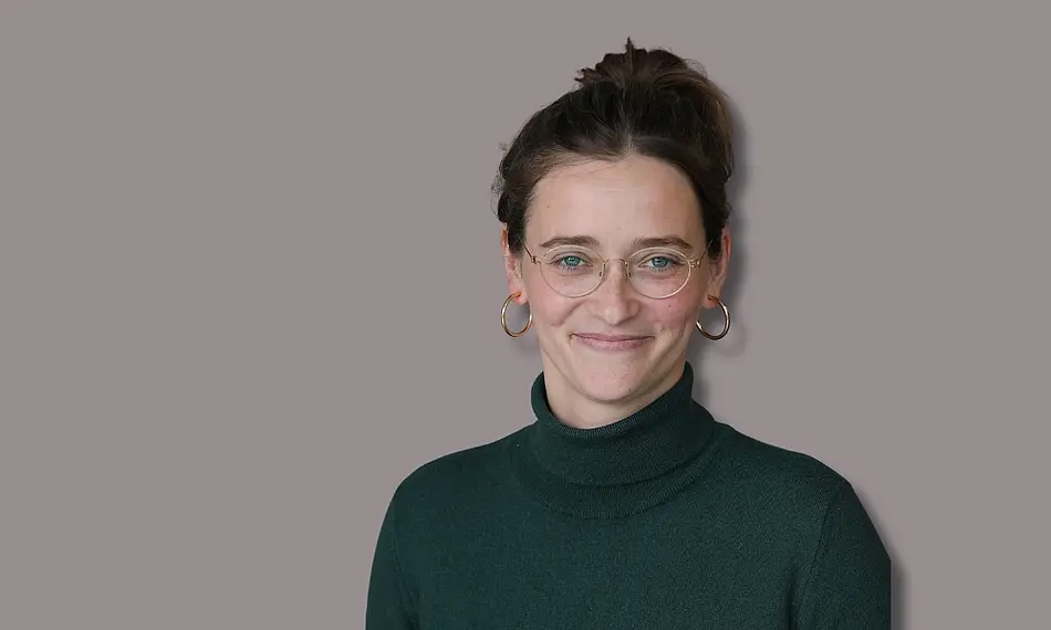 Porträtfoto von Lena Engel (lächelnd, weiße Frau mit dunklen langen Haaren hochgesteckt, dunkelgrüner Rollkragenpullover, runde Brille und goldene Ohrringe) vor grauem Hintergrund, angedeuteter Schatten