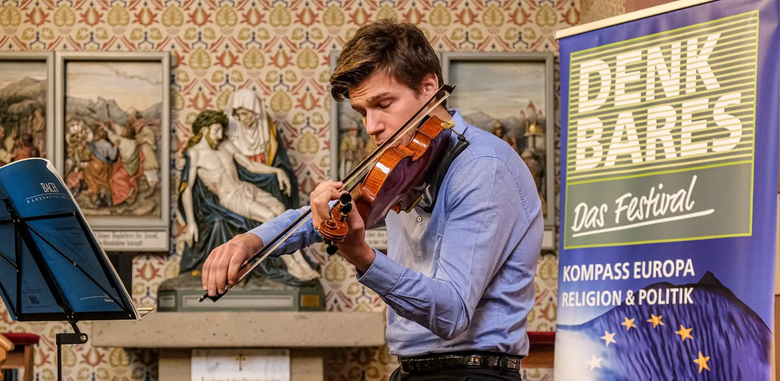 Ein junger Mann spielt Geige. Im Hintergrund sieht man einen Aussteller des Festivals
