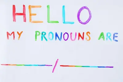 Person holding up a piece of paper that reads "Hello, my pronouns are" in rainbow colours