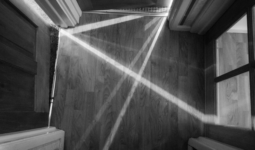 Schwarz-Weiß-Bild von sich kreuzenden Lichtstrahlen, die durch Türspalten in einen kleinen Flur mit Holzfußboden fallen