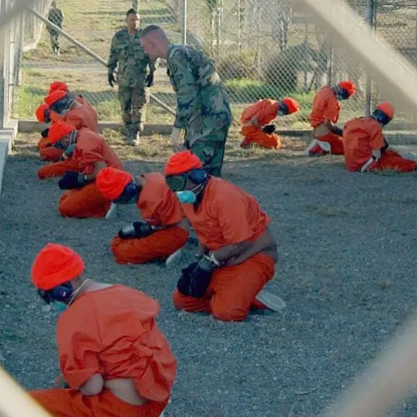 Detainees in orange jumpsuits sit in a holding area under the watchful eyes of Military Police at Camp X-Ray at Naval Base Guantanamo Bay, Cuba