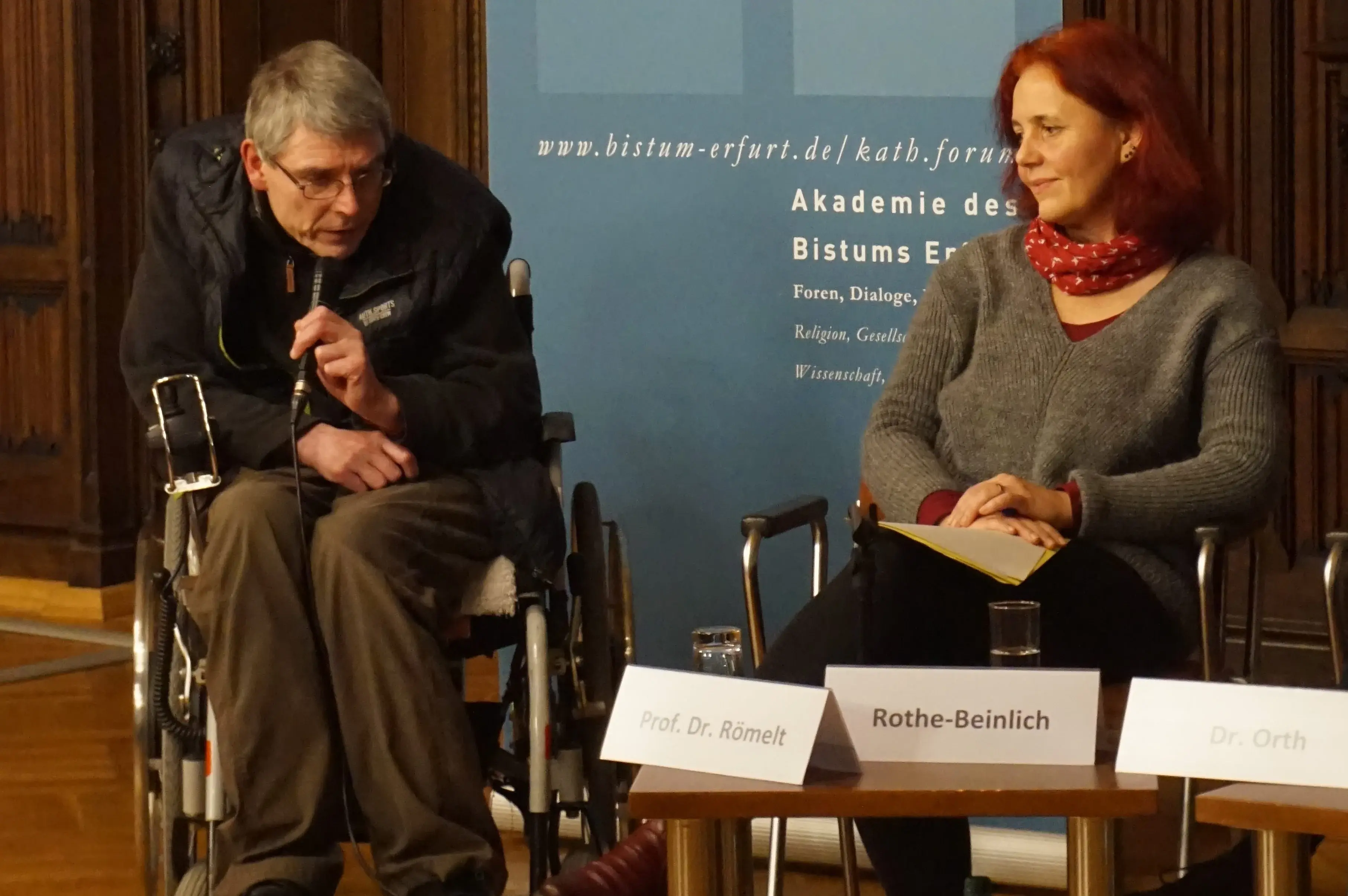 Prof. Dr. Josef Römelt spricht zum Publikum (mit Mikrofon; links) und Astrid Rothe-Beinlich (rechts) vor einem Roll Up Banner des Katholischen Forums im Land Thüringen