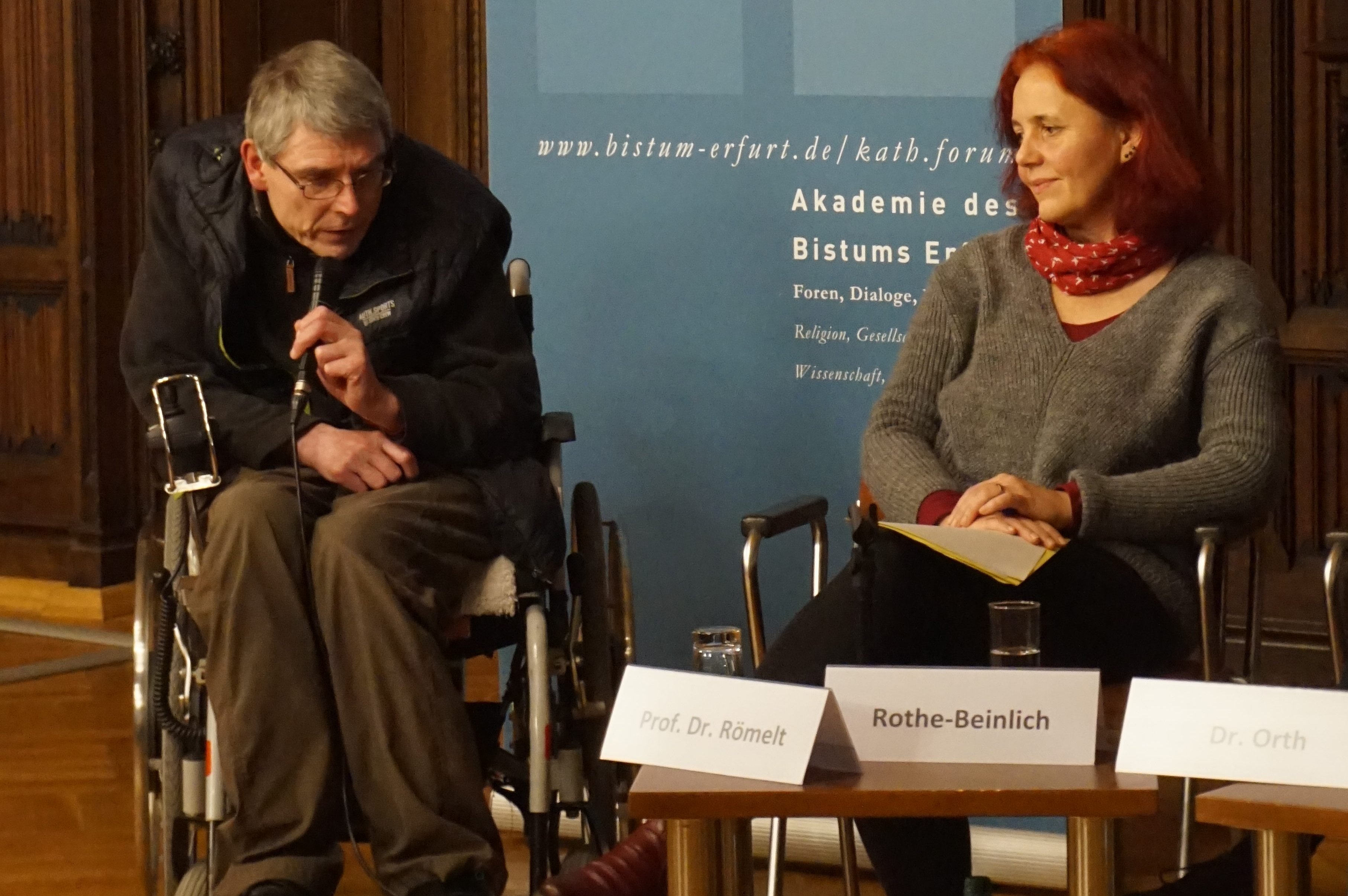 Prof. Dr. Josef Römelt spricht zum Publikum (mit Mikrofon; links) und Astrid Rothe-Beinlich (rechts) vor einem Roll Up Banner des Katholischen Forums im Land Thüringen
