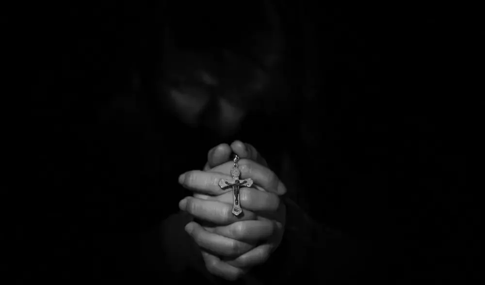 Betende Hände, die ein Kreuz halten - schwarzer Hintergrund (Symbolbild Vergebung)