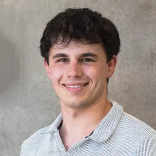 Porträtfoto von Kamil, internationaler Student aus Krakau, lächelnd in kamera, helles Hemd mit Kragen und schwarze, leicht lockige Haare, vor grauem Hintergrund