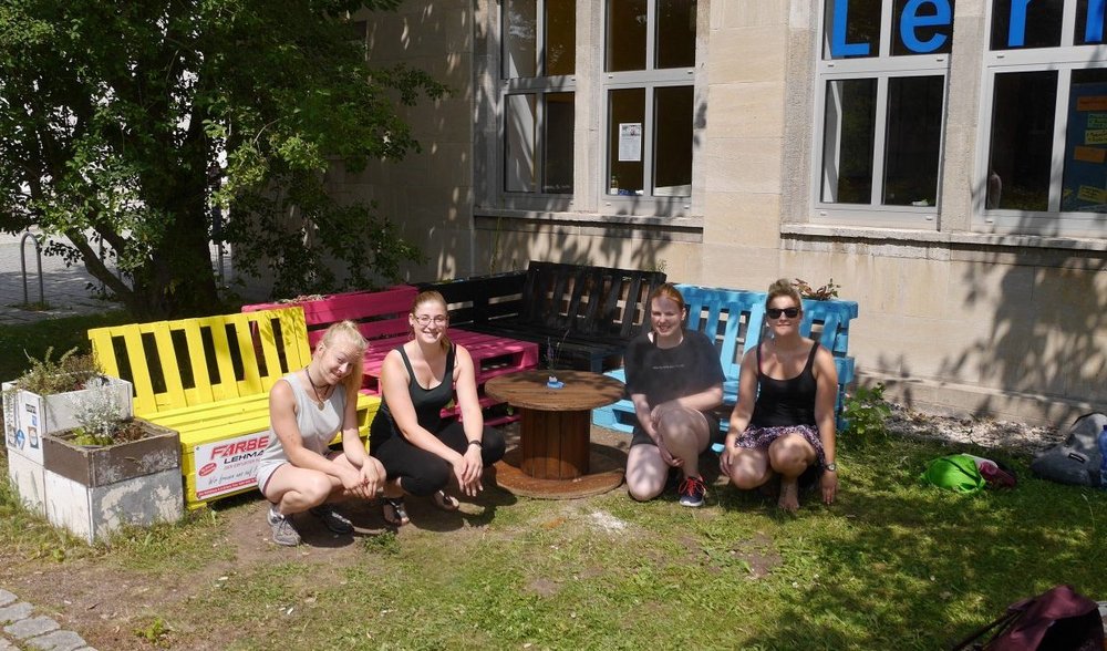 vier Studentinnen vor den neu bemalten Palettenmöbeln vor dem Gebäude der Lernwerkstatt