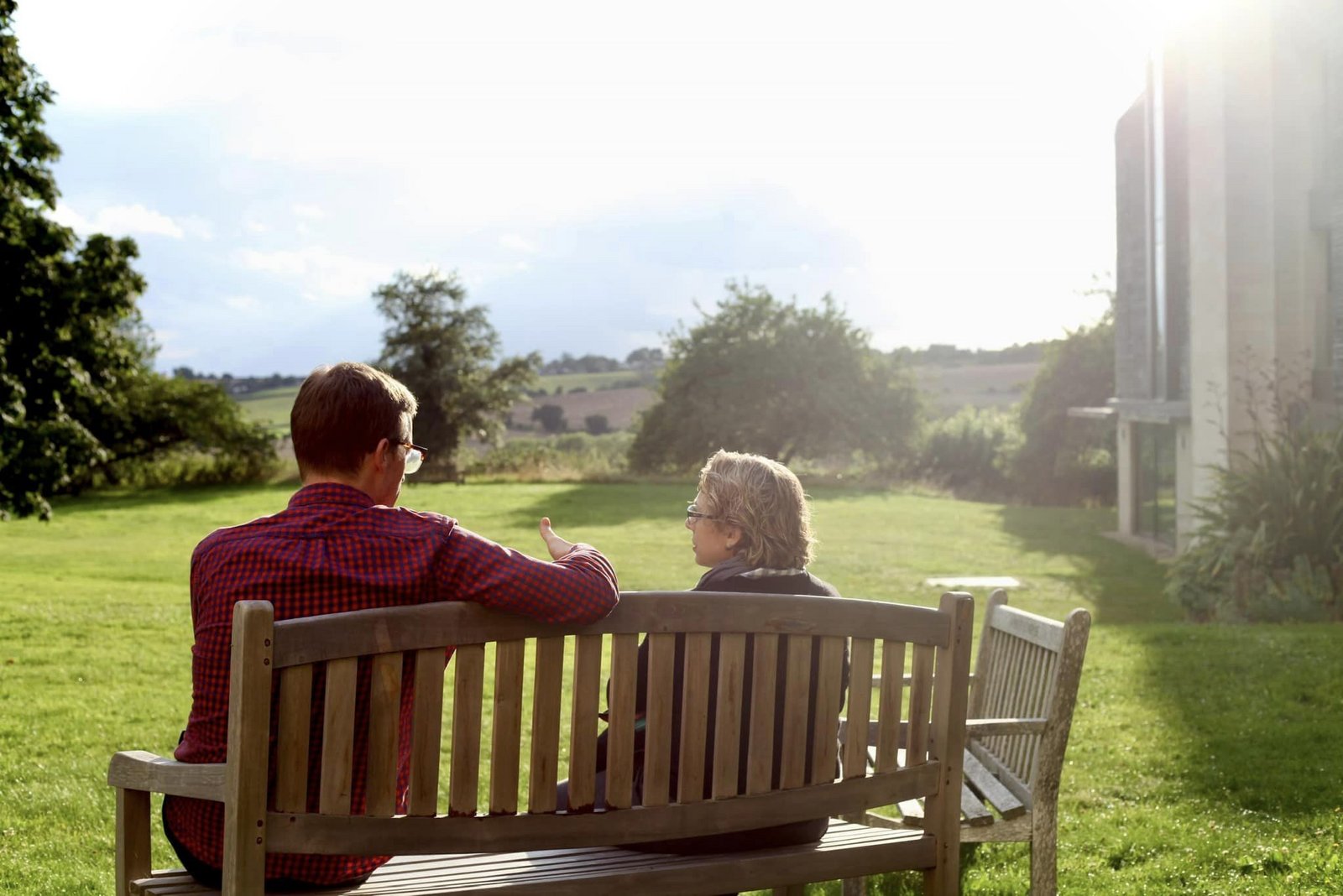 Ein Mann und eine Frau sind von hinten zu sehen, während sie auf einer Bank in der Sonne sitzen und miteinander diskutieren.