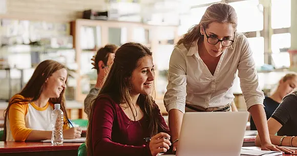 In einem Seminarraum eine Lehrende und Studierende schauen gemeinsam auf einen Laptop, im Gespräch