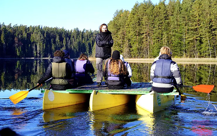 auf See Leute bei einer Kanufahrt, eine junge Frau steht mit dem Gesicht in die Kamera und erklärt etwas, im Hintergrund Tannenwald