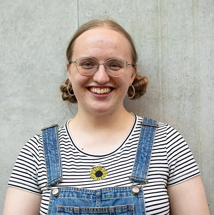 Weronika lächelnd mit Brille, zusammen gebundenen Haaren, Streifentop und Jeanslatzhose