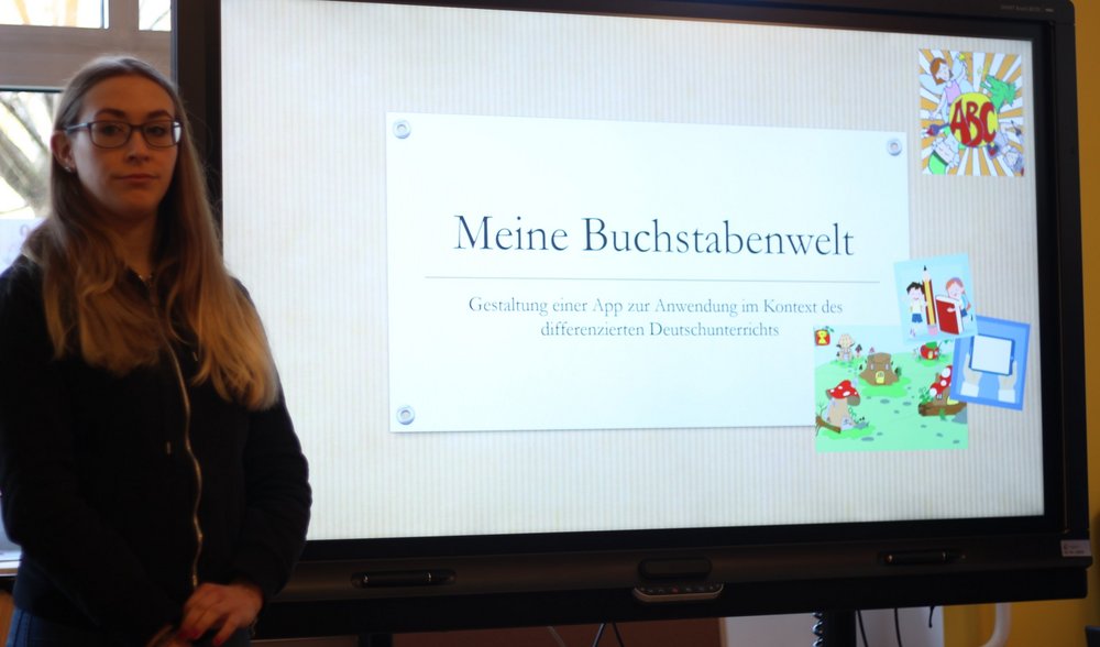 Studentin steht neben einem Smartboard und präsentiert ihr Projekt "Meine Buchstabenwelt - Gestaltung einer App zur Anwendung im Kontext des differenzierten Deutschunterrichts"