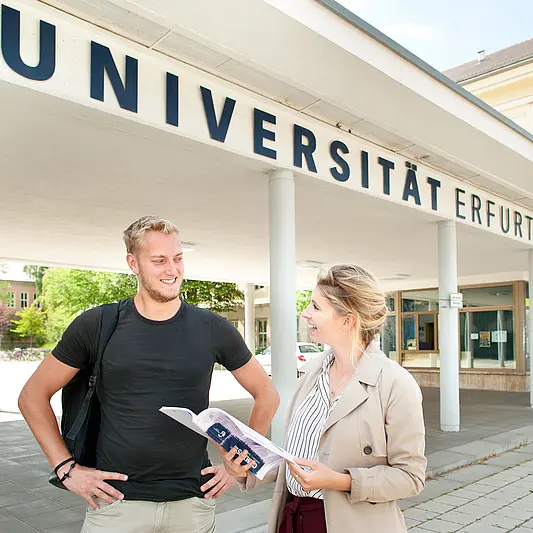 Ein Student und eine Studentin stehen vor dem Haupteingang der Universität Erfurt