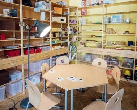 Blick in den Lernraum der Lernwerkstatt (Tisch mit Stühlen, dahinter Regale mit Materialien und Büchern)