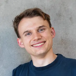 Porträtfoto von Max, schräg gehaltener Kopf, lächelnd in kamera, kurze dunkle Haare, trägt ein dunkelblaues T-Shirt vor grauem Hintergrund