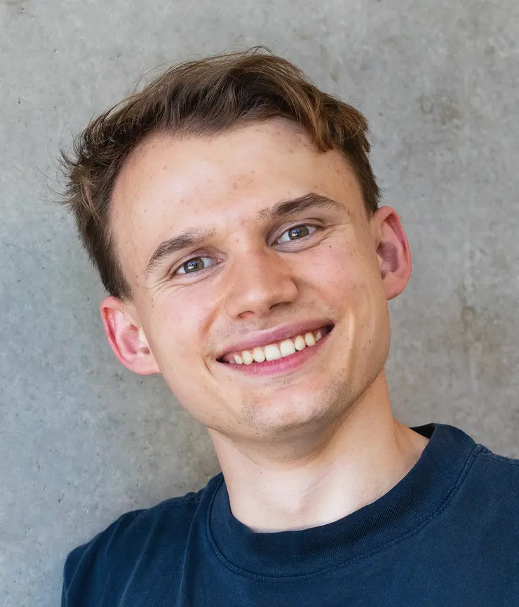 Porträtfoto von Max, schräg gehaltener Kopf, lächelnd in kamera, kurze dunkle Haare, trägt ein dunkelblaues T-Shirt vor grauem Hintergrund