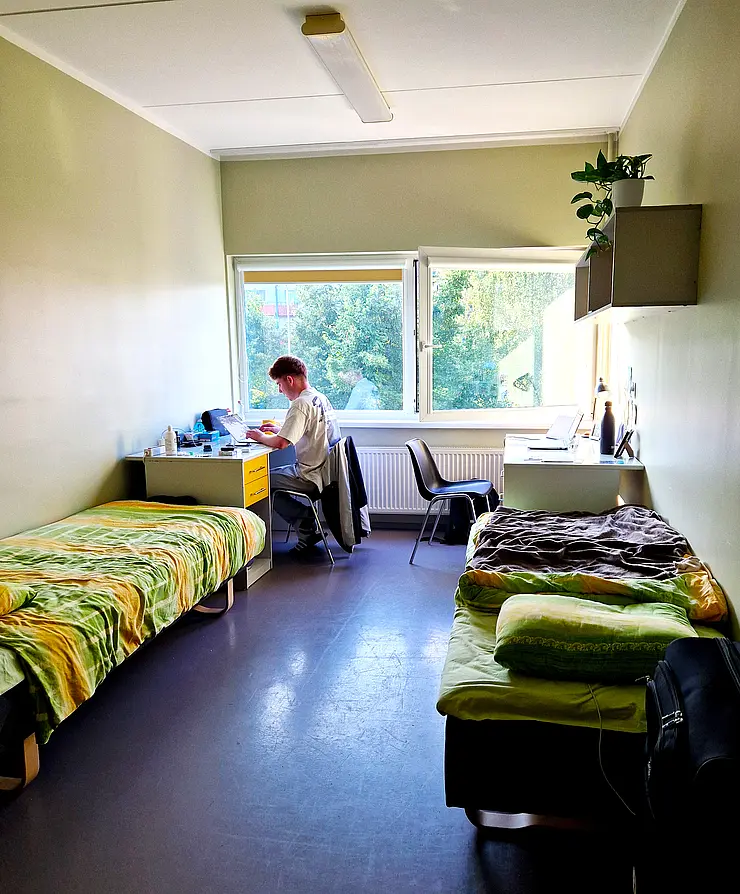 Foto aus dem Wohnheim in Tartu - Tobias sitzt am Schreibtisch mit Blick aus dem Fenster auf Bäume, im Vordergrund zwei Betten, das Zimmer wird von zwei Studierenden bewohnt