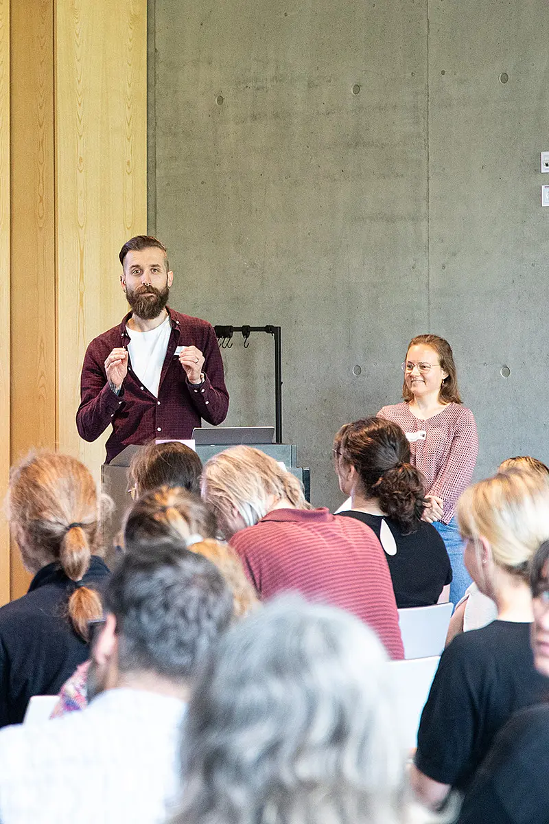 Zwei Vortragende vor einem Publikum, das mit dem Rücken zur Kamera sitzt