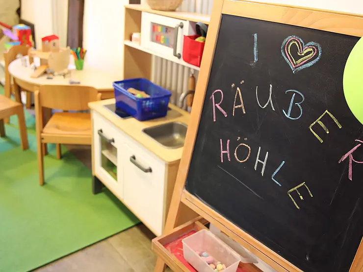Foto der Inneneinrichtung der Kinderbetreuung "Räuberhöhle": zu sehen eine Tafel, auf der mit bunten Stiften steht: I Herz Räuberhöhle, ein grüner Teppich, auf dem ein kleiner Küchenschrank und ein Tisch mit Spielzeug steht