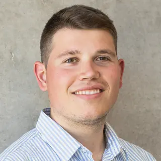 Porträtfoto von Elias, Student der Wirtschaftswissenschaft, freundlich lächelnd, kurze dunkelblonde Haare, hellblaues Hemd vor grauem Hintergrund