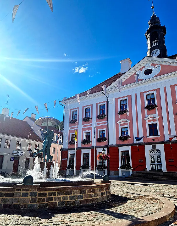 weiß-rotes Rathaus von Tartu, blauer Himmel, im Vordergrund ein Brunnen, Sonnenschein