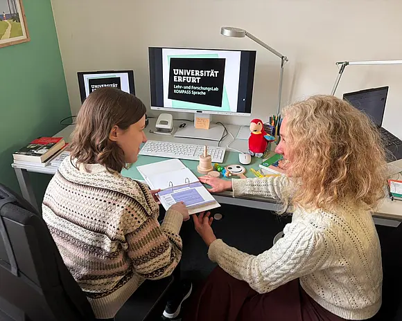 Zwei Kolleginnen sitzen nebeneinander an einem Schreibtisch und tauschen sich über Fördermaterial aus, was auf dem Tisch liegt. 