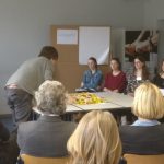 Teilnehmer:innen eines Workshops stehen im Kreis um einen Tisch mit kleinen Plastiklöffeln und ordnen diese unterschiedlich an