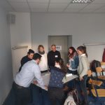 Teilnehmer:innen eines Workshops stehen im Kreis um einen Tisch herum und besprechen in Gruppenarbeit eine Aufgabe des Workshops im Rahmen der 11. internationalen Lernwerkstättentagung in Erfurt